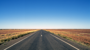 baner zdjecie szosa asfaltowa ciagnaca sie w przod zdjecie zrobone na srodu szowy szosa ciagnie sie po horyzont teren wokol szosy to niezabudowane terany wypalona trawa niebo blekitne bez chmur